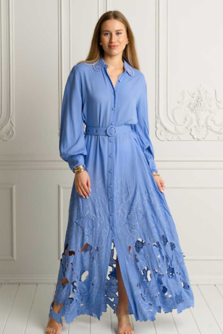 A woman wearing a light blue long-sleeve maxi shirt dress with a matching belt and lace-up front, standing in a white paneled room.