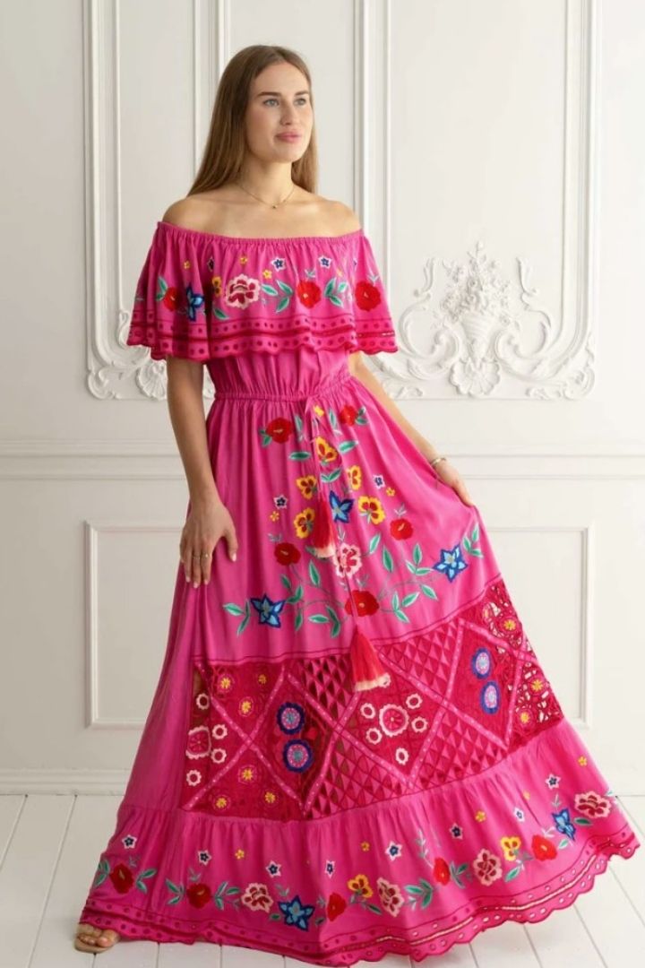 Woman wearing a pink off-shoulder dress with colorful embroidery against a white wall.