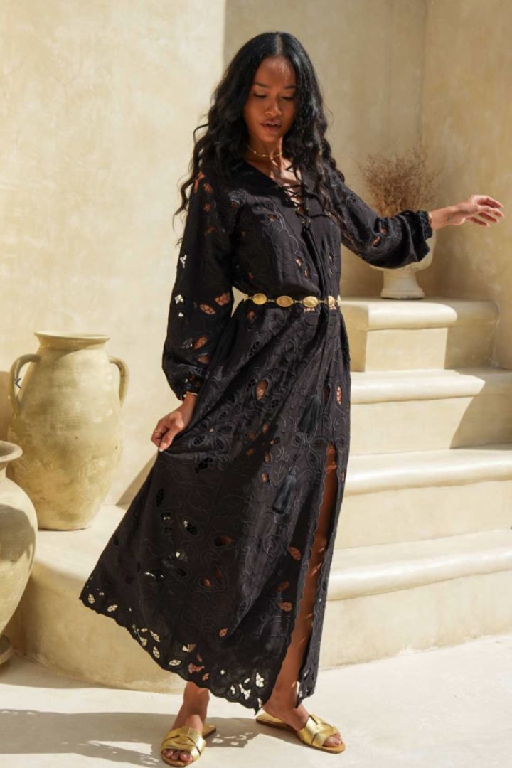 A woman wearing a black long-sleeve bohemian maxi dress with distressed cutouts and a gold belt, standing in front of beige stone steps and clay pots.