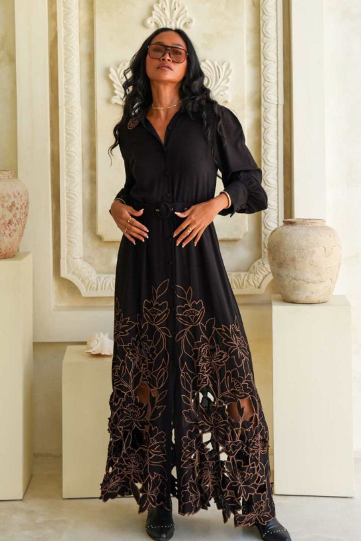 A woman modeling a black long-sleeve maxi dress with a sheer floral lace overlay concentrated along the hem, standing in an elegant interior setting with decorative vases.