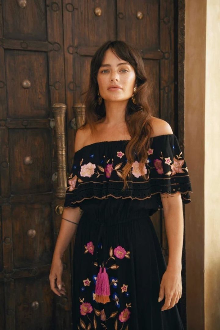 Woman wearing a black off-shoulder dress with floral embroidery in front of a wooden door.