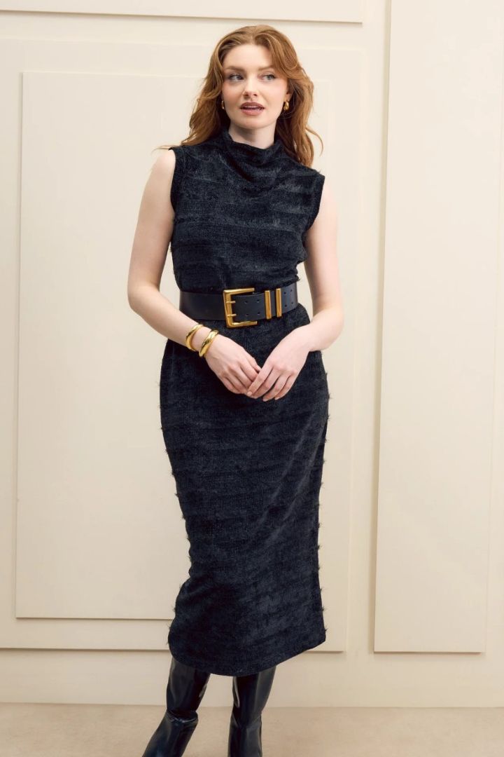 Woman wearing a black dress with a gold belt against a beige wall.