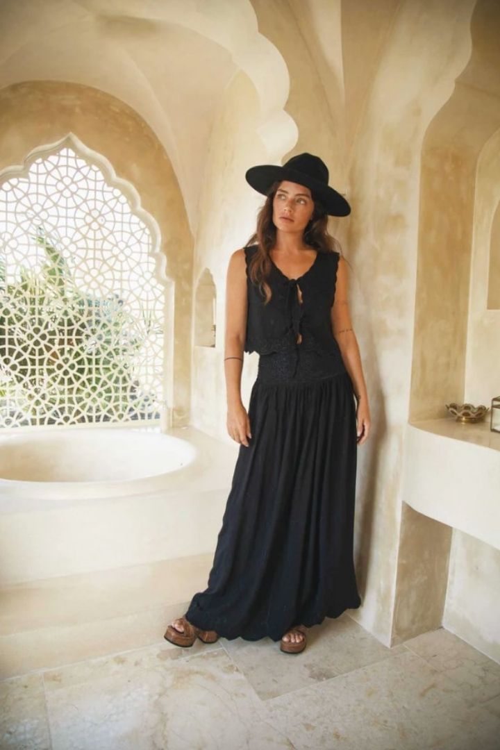 Woman in a black top, skirt and hat standing in an arched stone hallway.