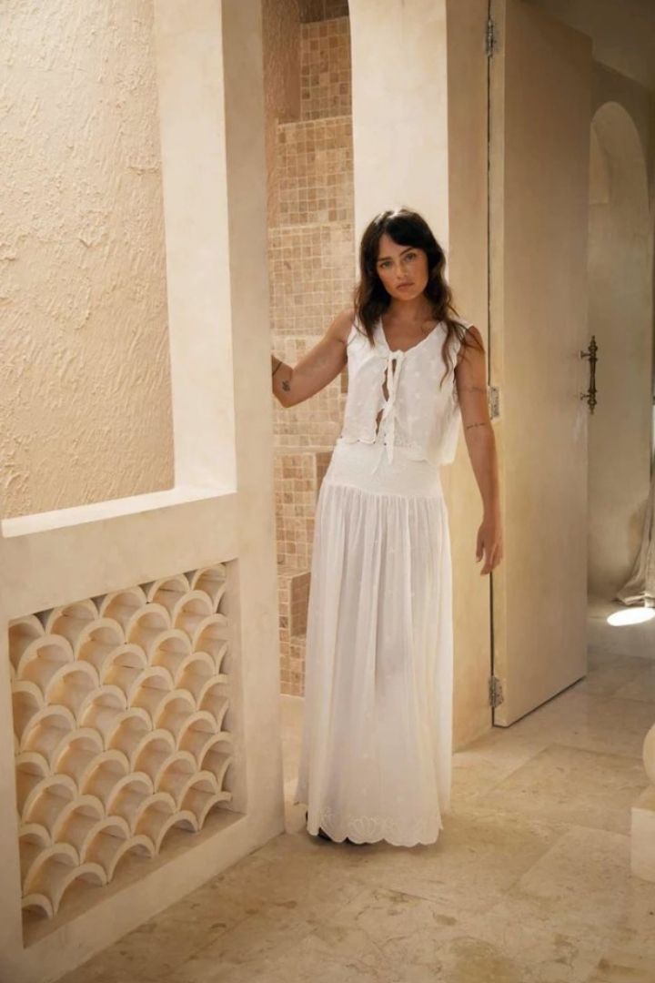 Woman in a white top and maxi skirt standing in a sunlit room with architectural columns.