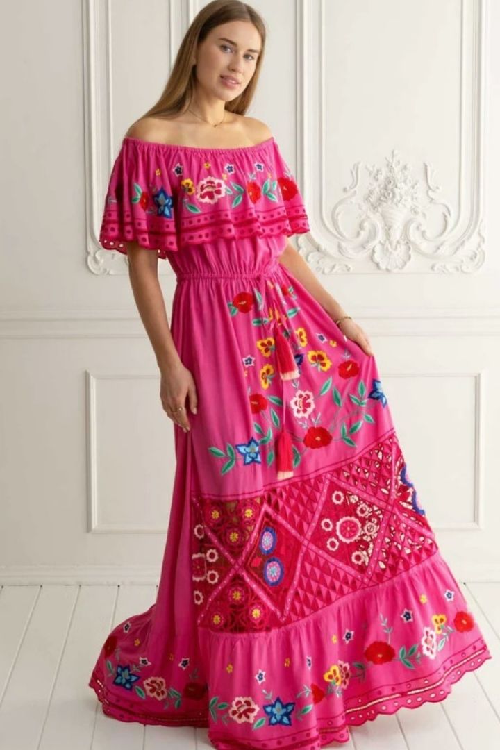 Woman wearing a pink off-shoulder dress with floral embroidery against a white wall.