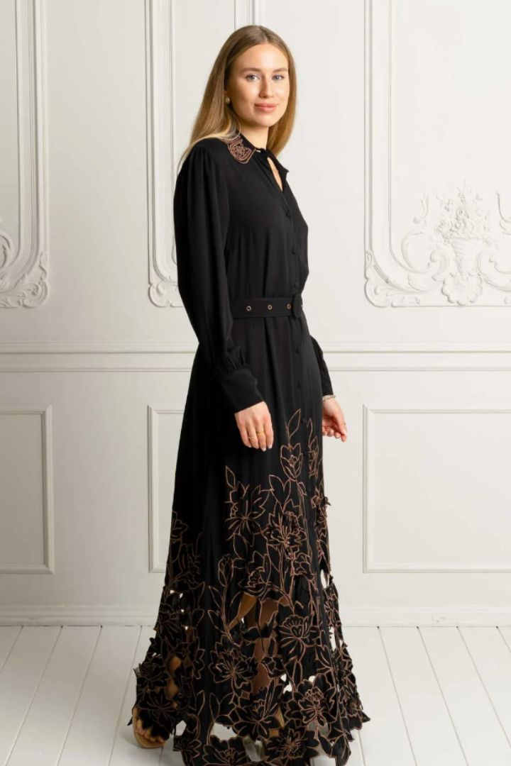 A woman modeling a long black dress with brown floral embroidery and lace details, standing against a white paneled wall.