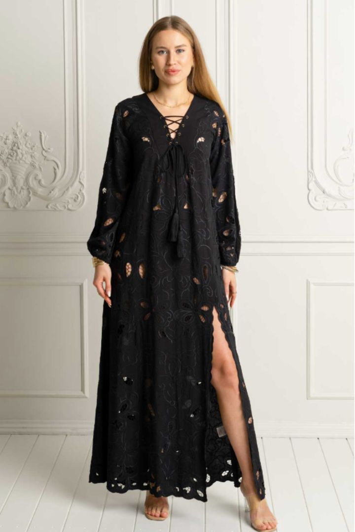A woman modeling a long black lace maxi dress with a tie-front neckline, lace-up detail at the chest, and a high side slit, standing against a white paneled wall.