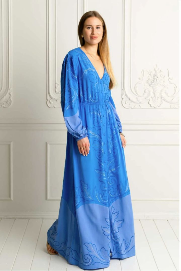 Woman wearing a blue embroidered long sleeve dress with buttons against a white wall with a side view of the dress