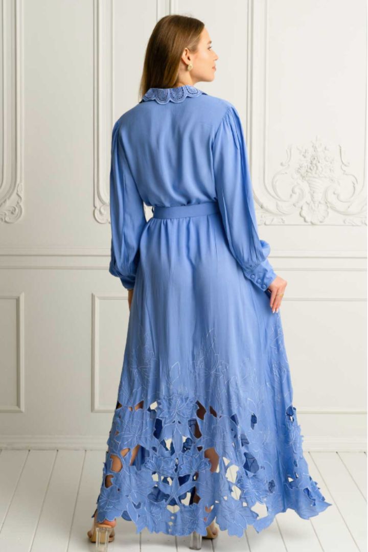 A woman wearing a long-sleeved blue maxi dress with a ruffled collar, belted waist, and intricate floral lace cutout detailing along the hem, shown from the back against a white paneled wall.