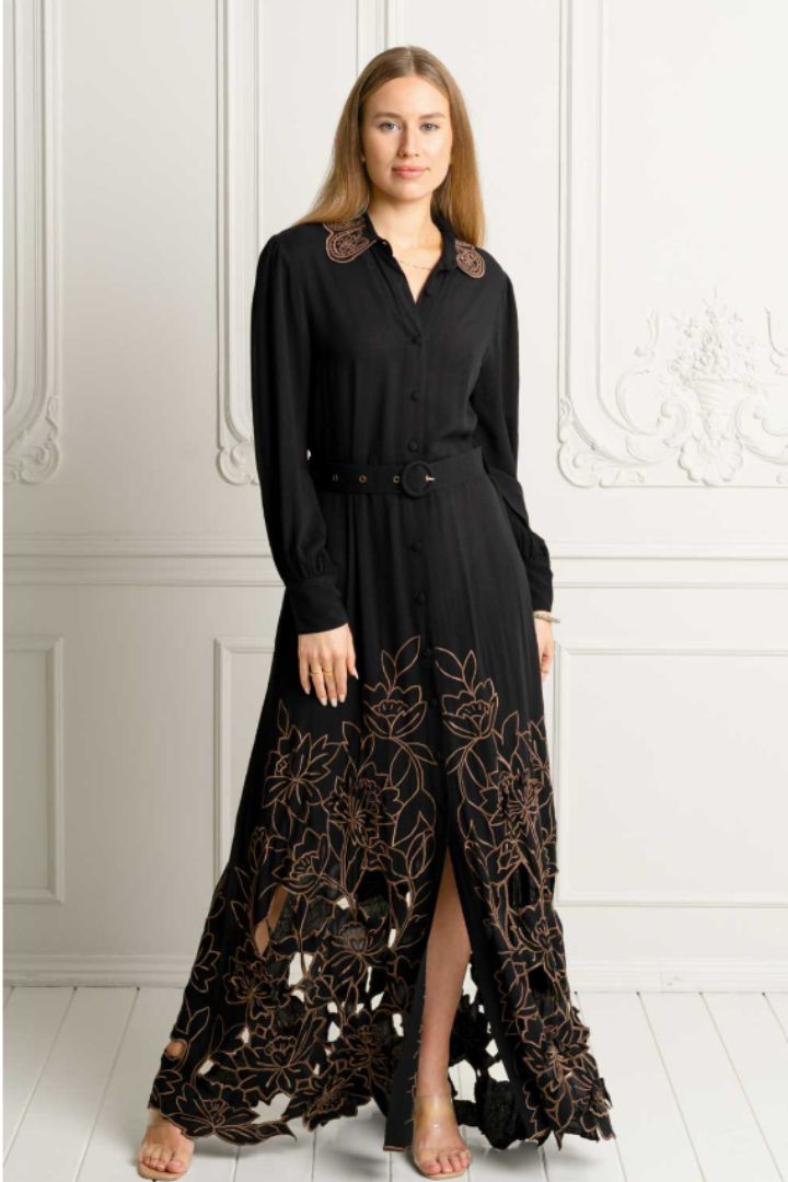 A woman modeling a long black maxi dress with long sleeves, a button-front bodice, a thin waist belt, and a dramatic sheer floral lace hem with cutouts, standing against a white paneled wall.