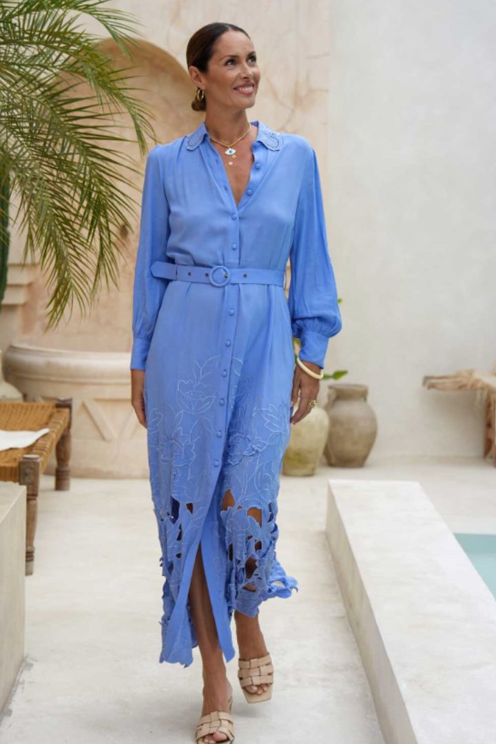 A woman wearing a light blue long-sleeve midi dress with a matching belt and distressed lace-up hem, walking in a sunlit outdoor courtyard.