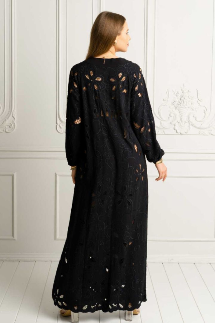 A woman viewed from the back wearing a long black embroidered dress with sheer cutout details, standing against a white paneled wall.