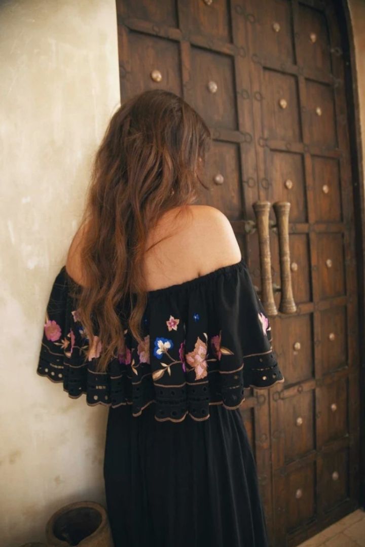 Woman wearing a black off-shoulder dress with floral embroidery in front of a wooden door.