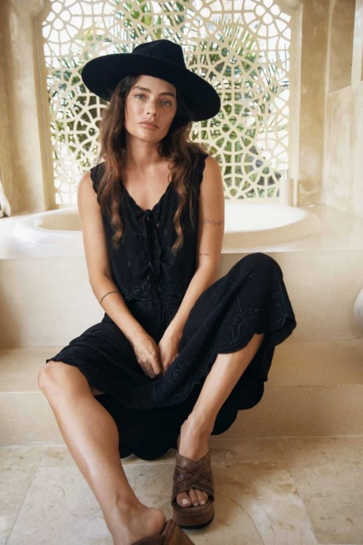 Woman in a black skirt, top and hat sitting on a stone floor with a decorative wall in the background