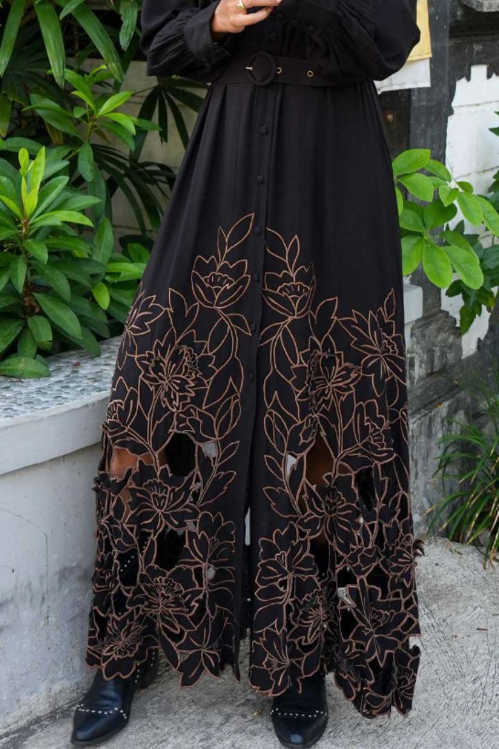 A black maxi dress with large floral embroidery and cutout details in a warm brown tone, worn by a model standing outdoors on a stone path with green plants in the background.