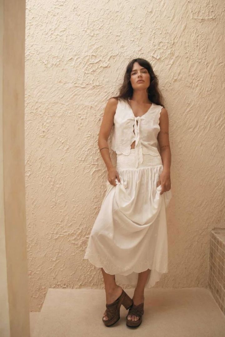 Woman wearing a white skirt and top standing against a textured beige wall.