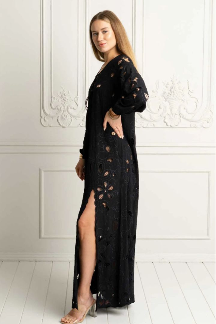 A woman modeling a long black open-knit maxi dress with floral lace cutouts and a thigh-high slit, standing against a white paneled wall.