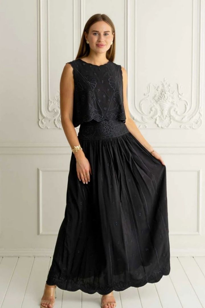 Woman wearing a black lace top and skirt standing against a white wall.