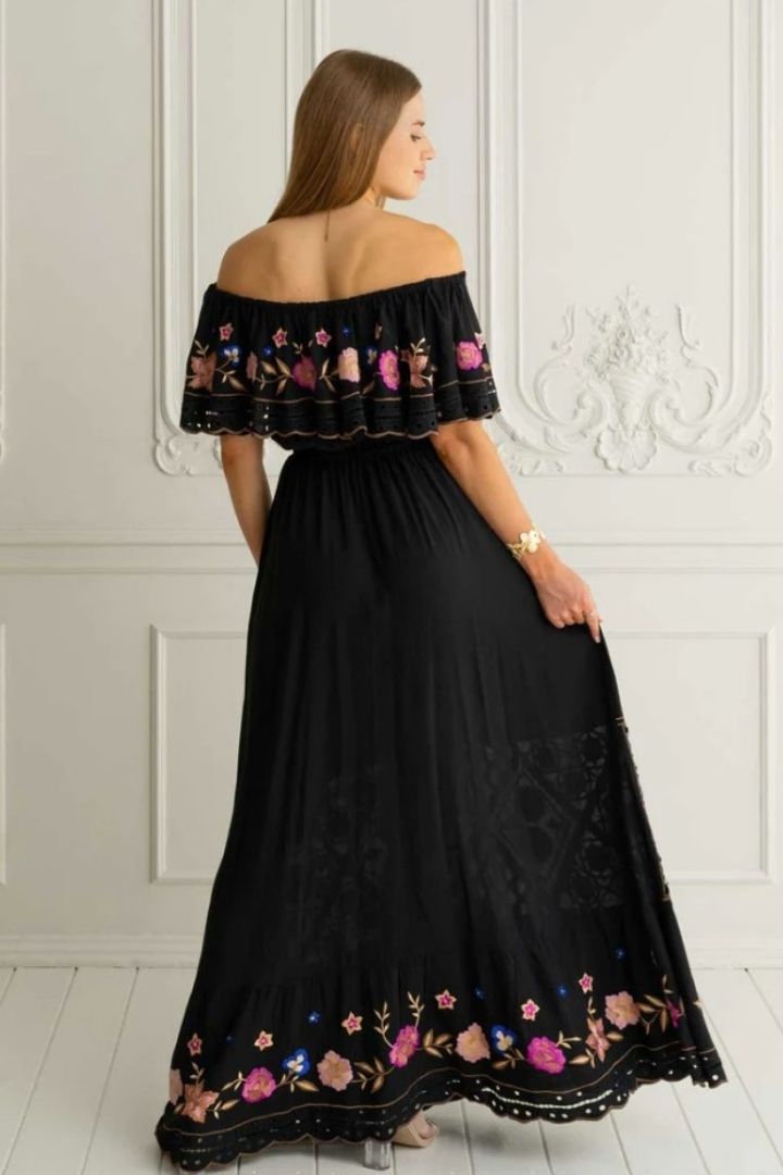 Woman wearing a black off-shoulder dress with floral embroidery in a white room.