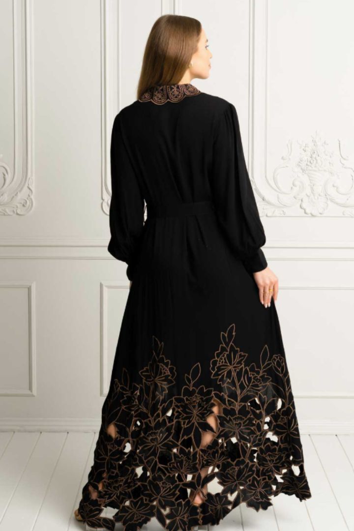 A woman viewed from the back wearing a long black dress with a high lace collar and a large floral lace overlay near the hem, standing against a white paneled wall.