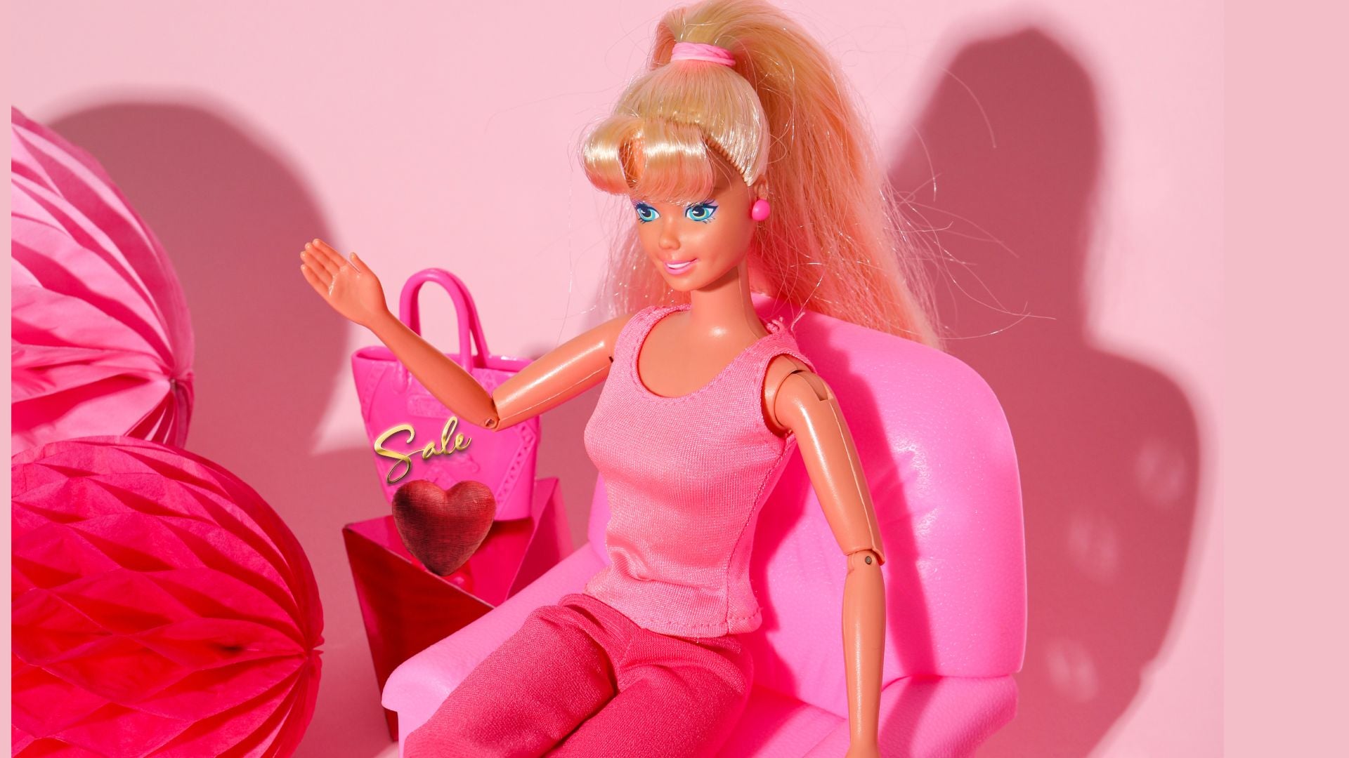 Barbie doll sitting on a pink chair with a pink handbag next to her against a pink background
