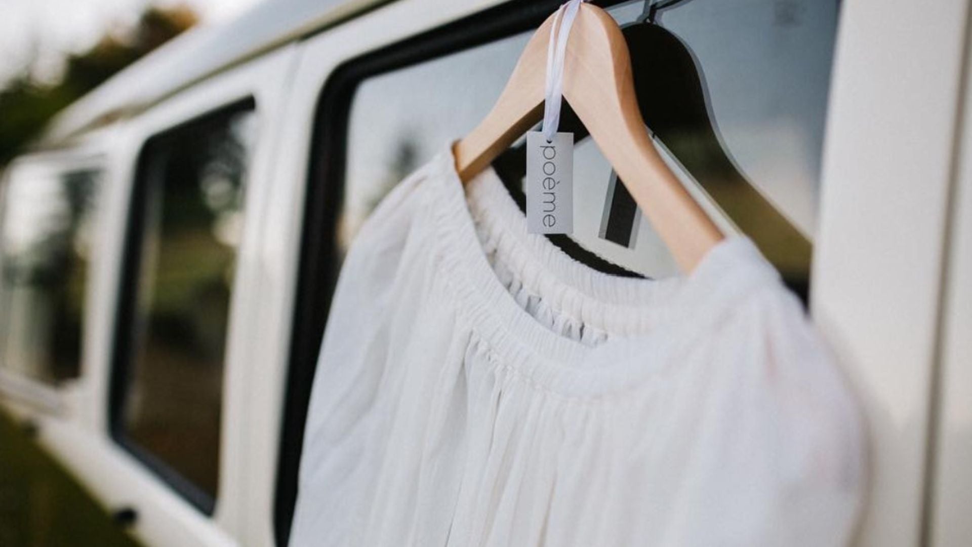 A white textured blouse hanging on a wooden hanger with a 'Poeme' tag, positioned in front of a white vehicle window.