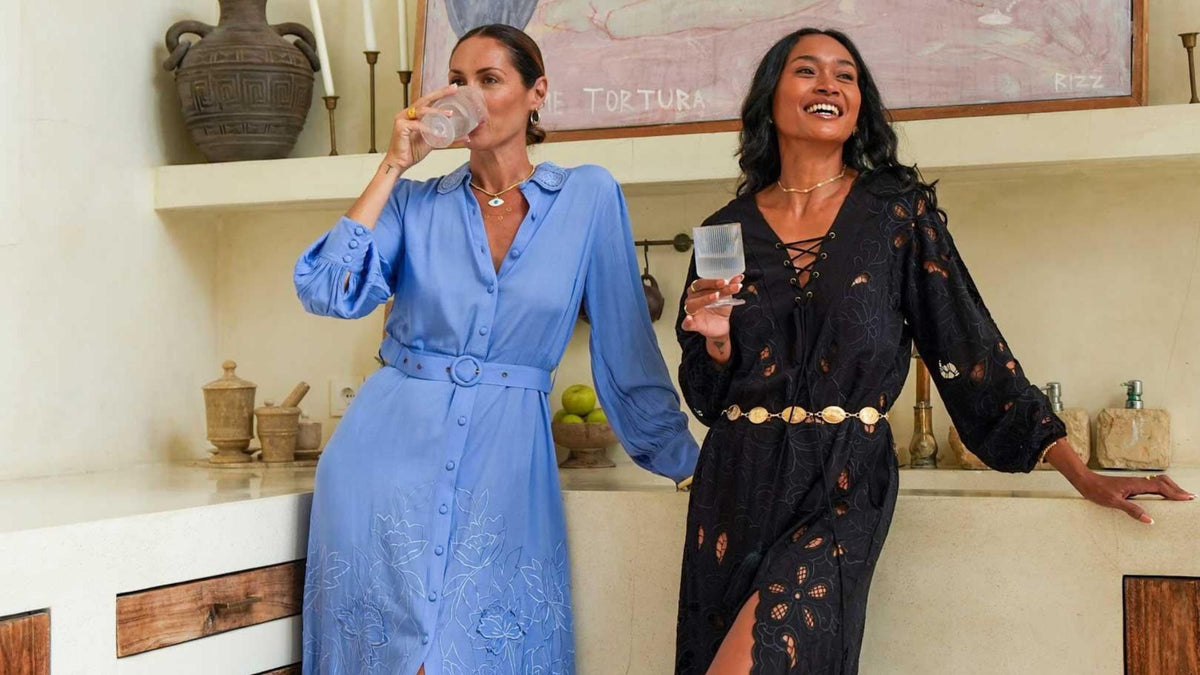 Two women, one wearing a long blue embroidered belted maxi dress sipping water and the other smiling in a black cut out dress with a gold belt holding a glass of water